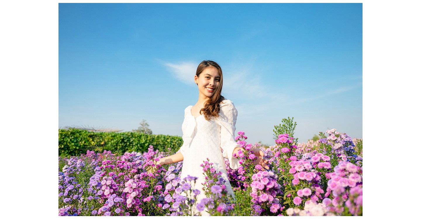 Eine Frau in einem weißen Kleid steht draußen zwischen vielen Blumen und posiert für ein Foto.

